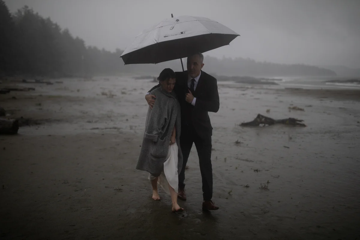 Un couple s'abrite sous un grand parapluie sur une plage brumeuse, sous la pluie (Originale)