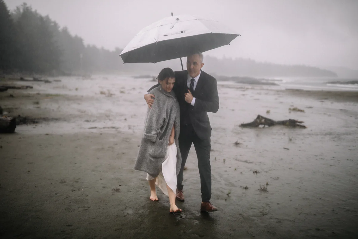 Un couple s'abrite sous un grand parapluie sur une plage brumeuse, sous la pluie (Retouche PHAiTO)