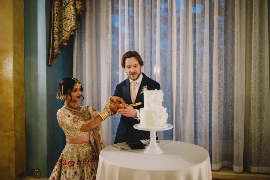 Le couple coupe un gâteau de mariage blanc devant une fenêtre aux voilages bleutés