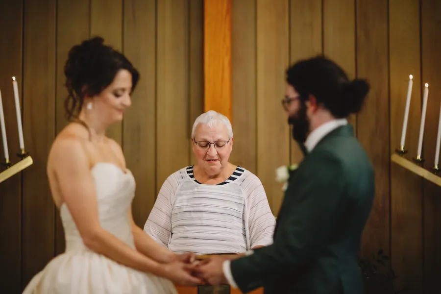 La mariée sourit, les yeux baissés, tandis que le couple se tient la main devant l'officiante