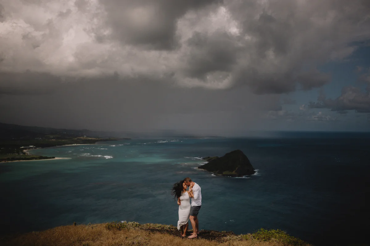Un couple s'embrasse au sommet d'une falaise herbeuse au-dessus d'une baie turquoise, sous un ciel gris et lourd