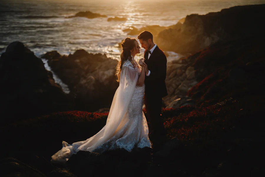 Un couple sur des rochers côtiers au coucher du soleil, le soleil irradiant sur l'océan derrière eux
