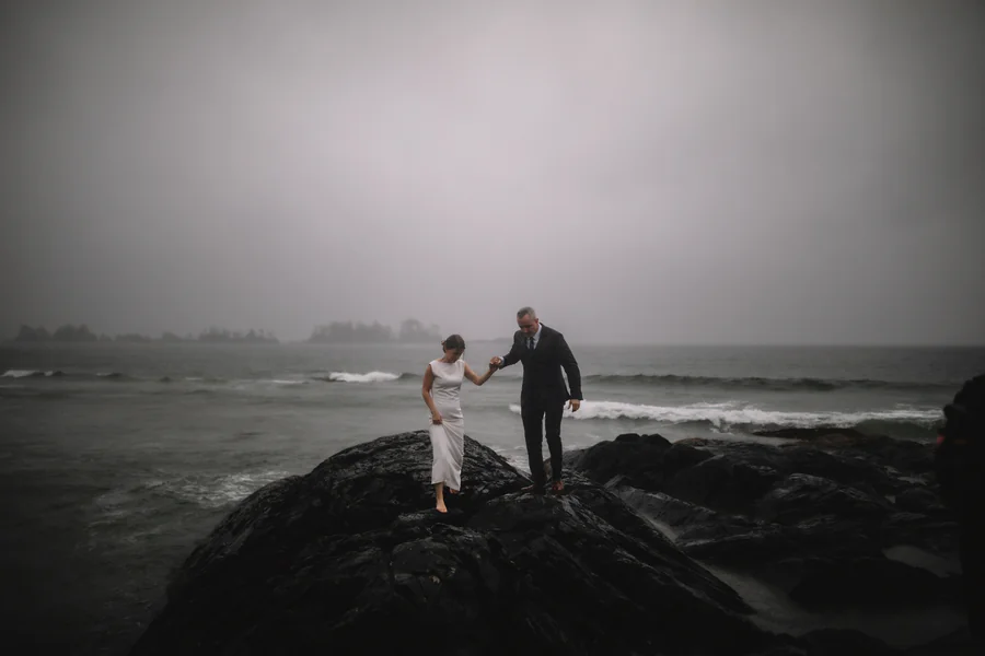 Un couple sur un rocher sombre, sur une plage brumeuse, le ressac pâle derrière eux