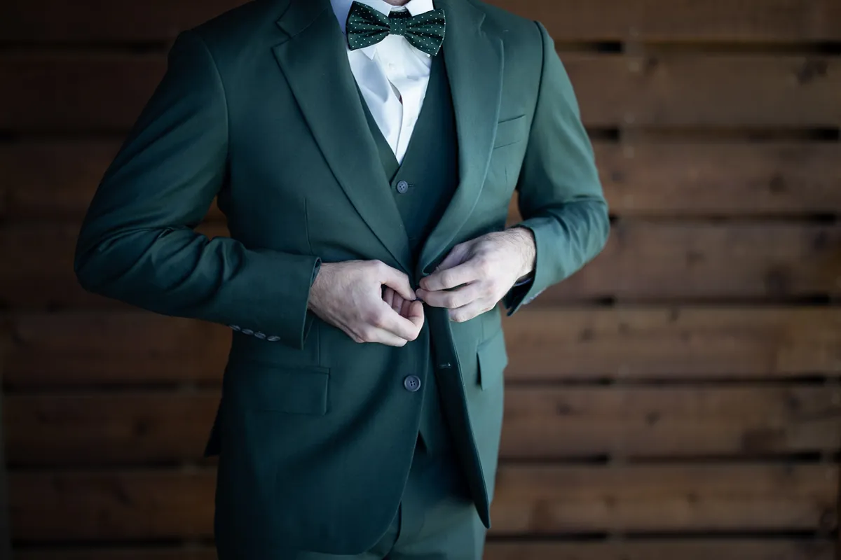Le marié boutonne la veste de son complet vert devant un mur en bois (Originale)