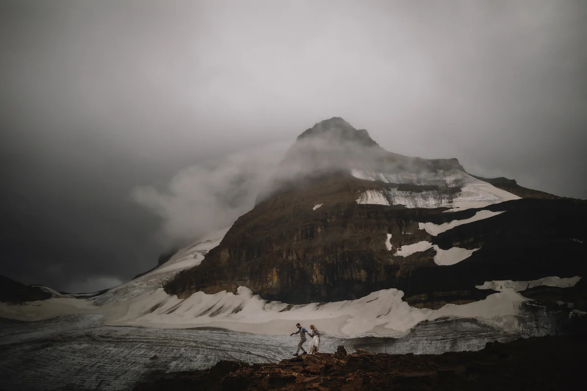 Un couple traverse un glacier sous un sommet enneigé enveloppé de nuages