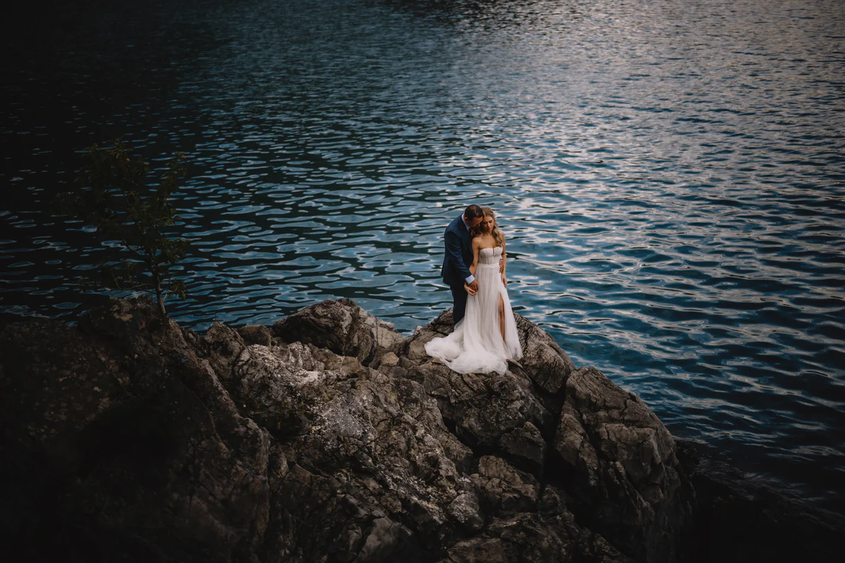 Un couple sur une corniche rocheuse au bord d'un lac d'un bleu profond, l'eau captant les dernières lueurs