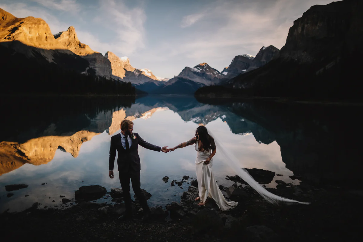 Un couple se tient par la main sur la rive rocheuse d'un lac de montagne immobile, les sommets dorés se reflétant dans l'eau