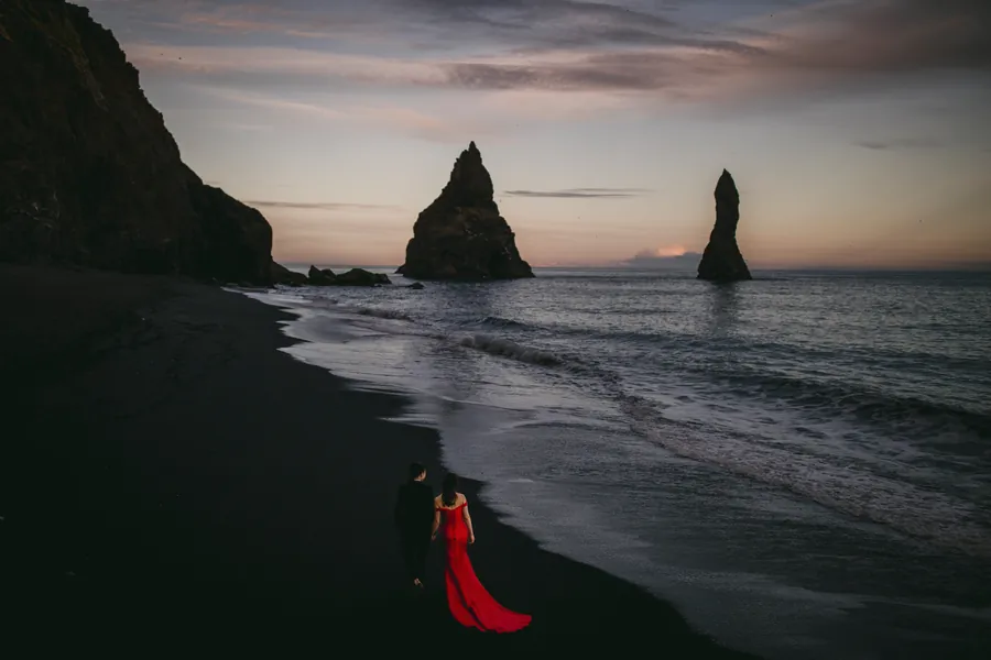 Femme en robe rouge sur une plage de sable noir, au pied d'aiguilles rocheuses