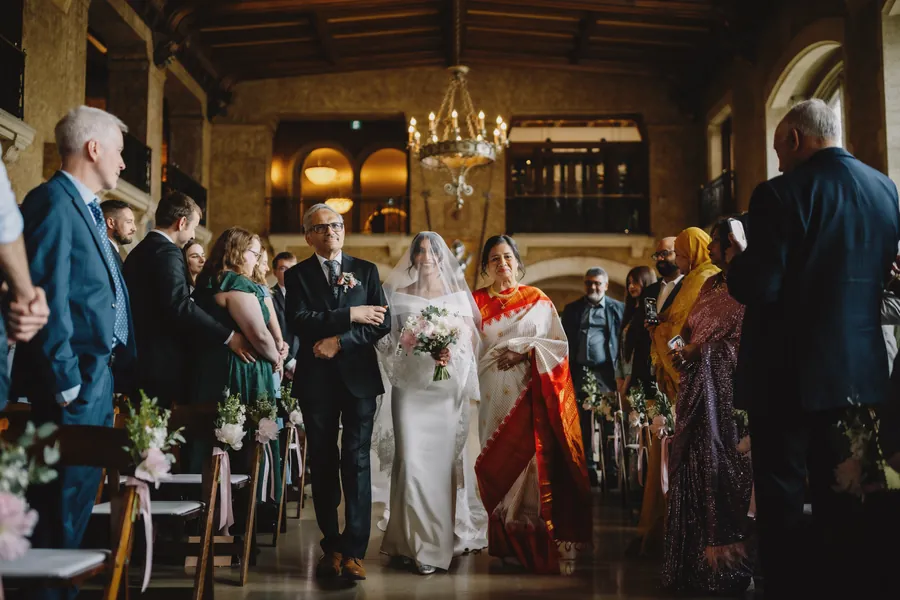 La mariée remonte l'allée avec sa famille sous les lustres d'une salle en pierre