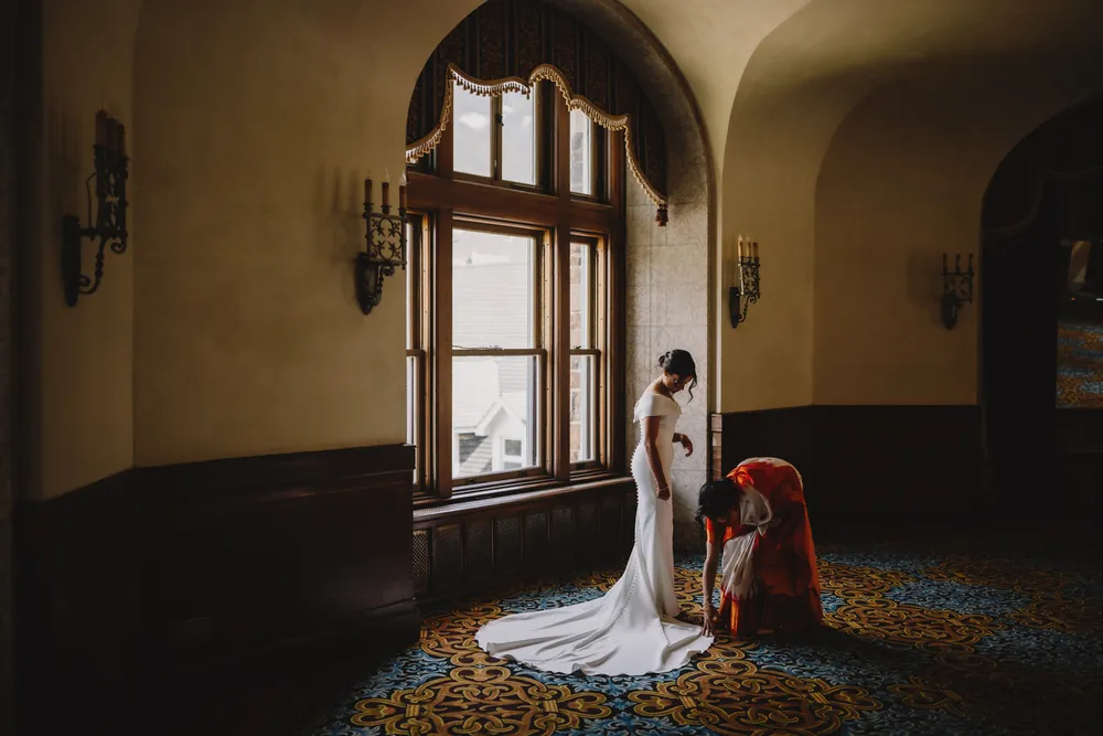 Mariée en robe de satin éclairée par une haute fenêtre cintrée, pendant qu'une invitée ajuste sa traîne