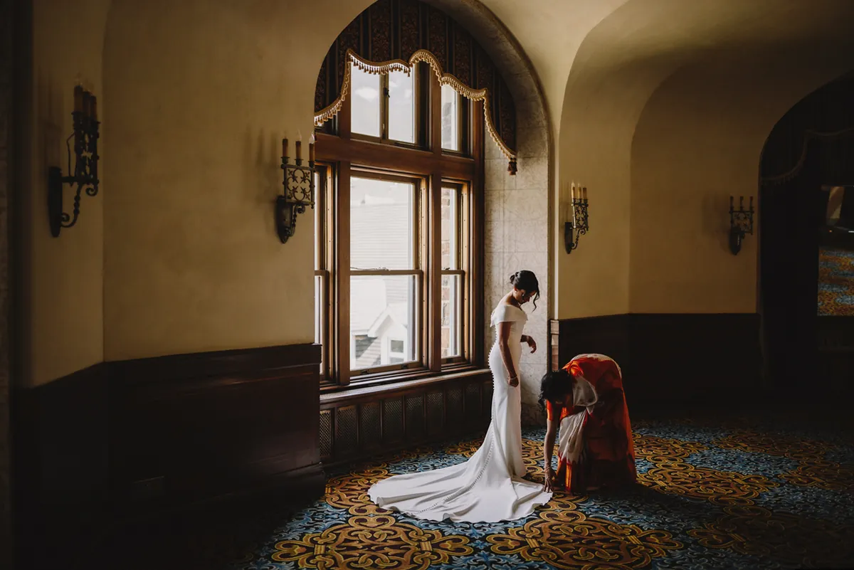 Mariée en robe de satin éclairée par une haute fenêtre cintrée, pendant qu'une invitée ajuste sa traîne